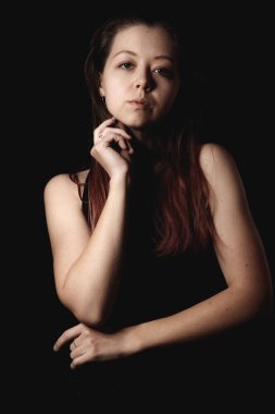 Uzun kahverengi saçlı, siyah bluzlu, eli çenesinde koyu bir arka plan kitap kapağına yaslanmış genç bir kadının stüdyo portresi.