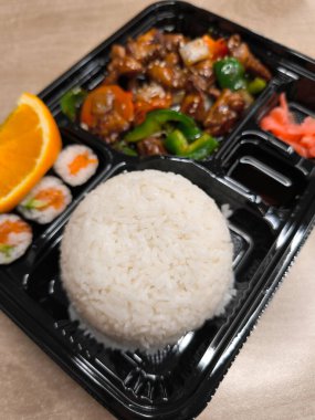 Buğulanmış beyaz pirinç, teriyaki tavuğu ve suşi zarafeti ferahlatıcı bir portakal dilimiyle birlikte bir bento kutusu, dengeli ve lezzetli bir öğle yemeği, fast food sunuyor.