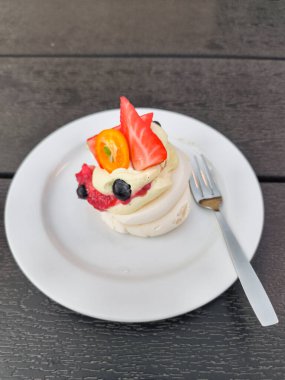 Çilek, kumkat, yaban mersini ve kremayla kaplı Pavlova pastası Prag 'daki koyu ahşap bir masada beyaz bir tabakta dinleniyor.