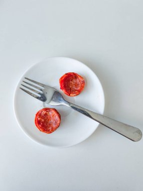 İki dilim pepperoni bir çatalla birlikte küçük beyaz bir tabakta oturur ve basit ama ilgi çekici bir mutfak sahnesinin fotokopisini çeker.