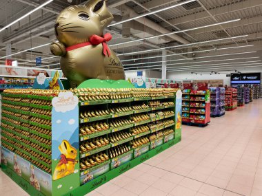 Prag, Çek Cumhuriyeti - 26 Mart 2025: Parlak ışıklı bir süpermarket koridorunda Lindt altın tavşanlarının canlı bir gösterisini koruyan dev şişirilebilir paskalya tavşanı