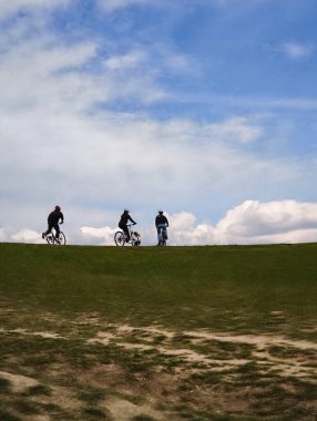 Üç bisikletçi bulutlu bir gökyüzünün altındaki yeşil bir çayırda bisikletlerini sürüyorlar. Açık hava aktivitelerinin ve sağlıklı yaşam tarzlarının tadını çıkarıyorlar.
