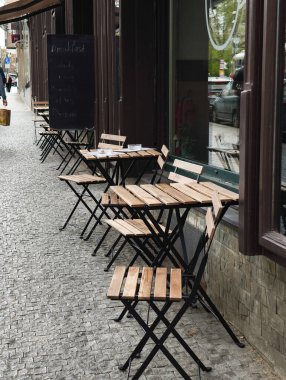 Kapalı bir restoranın önündeki kaldırımda duran ahşap masa ve sandalyeler huzurlu bir şehir manzarası yaratıyor.