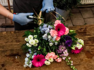 Çiçekçi rengarenk çiçeklerle dolu bir buket üzerinde çalışıyor, rafya kullanarak onları ahşap bir masaya bağlıyor.