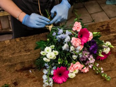 Mavi eldiven giyen çiçekçi ahşap bir çalışma masasında rafya ile renkli bir çiçek buketi bağlıyor.