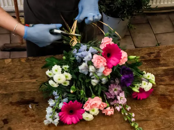 Çiçekçi rengarenk çiçeklerle dolu bir buket üzerinde çalışıyor, rafya kullanarak onları ahşap bir masaya bağlıyor.