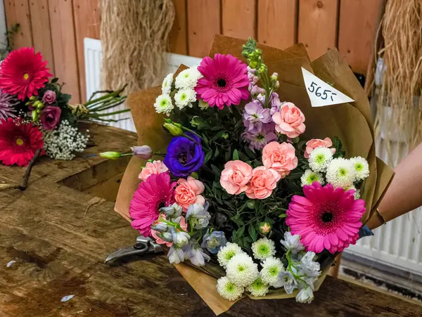 Kahverengi kağıda ve fiyata sarılmış gerberalar, karanfiller ve kasımpatılarla canlı bir buket hazırlayan çiçekçi.