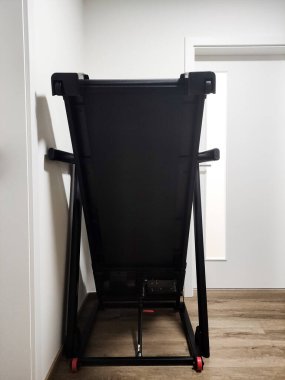 Çağdaş bir odada duvara yaslanmış katlanmış siyah koşu bandı. Ahşap döşeme ve bir kapı var. Uzayın verimli kullanımı ve modern tasarım estetiği sergileniyor.