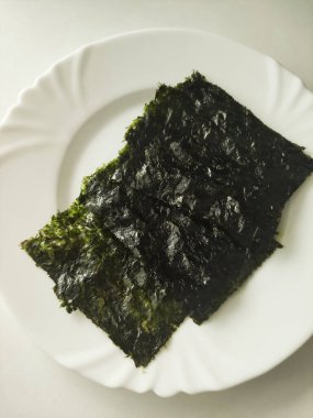 Pürüzsüz beyaz bir tabakta sergilenen koyu yeşil yosun parçacıkları, parlak yüzeyini ve katmanlı dokusunu vurguluyor, çeşitli yemeklerde aşçılık kullanımı için mükemmel, yemeklere lezzet ve besin katıyor.
