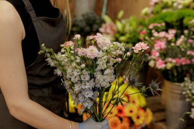 Çiçekçi dikkatlice bir buket rengarenk tarla kuşu, karanfil ve eryngium yerleştirirken çiçekçi dükkanında çeşitli çiçek kovanlarıyla çevrili Anneler Günü