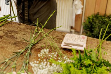Çiçekçi çiçek buketi hazırlıyor ve hesaplama maliyetlerini hesaplıyor. Etrafı çeşitli çiçek ve yapraklarla çevrili bir çiçekçide hesap makinesi kullanıyor.