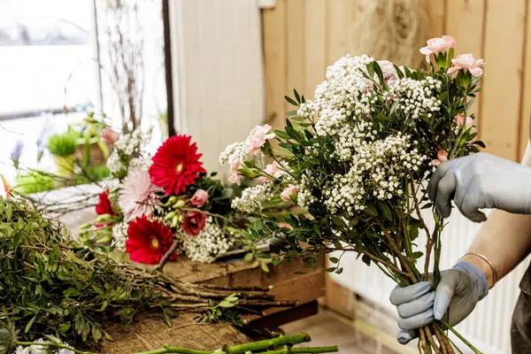 Çiçekçi, çiçekçi dükkanında bir buket pembe ve beyaz çiçekleri çingene ve yeşillikle birlikte bir ahşap masada düzenleyen eldiven giyiyor. Anneler Günü.