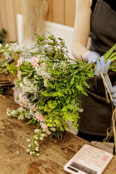 Eldiven takan bir çiçekçi yakınlarda hesap makinesi olan ahşap bir masada çiçek buketi düzenliyor. Anneler Günü.
