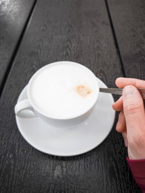 Sütlü kremalı kahve fincanı, kaşık tutan bir el, koyu ahşap bir masaya yerleştirilmiş, kahve sevenler ve hayranlar için samimi ve davetkar bir ortam yaratıyor. Boşluğu kopyala