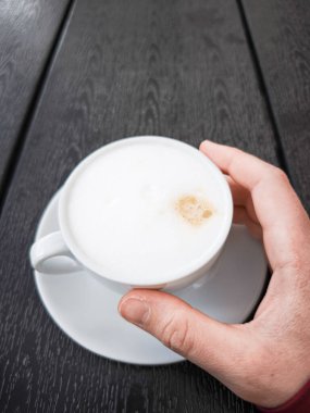 Bir elde tutulan kremalı kahve fincanı kapuçino koyu ahşap bir masanın üzerinde duruyor, nefis içecek deneyimi, kahve severler ve zengin lezzet ve dokulardan hoşlananlar için mükemmel. Boşluğu kopyala