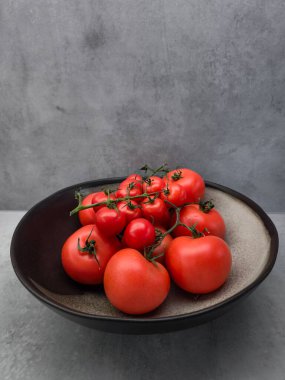Asmada olgun kırmızı domatesler ve vişneli domatesler, siyah bir kâsede sunuluyor, basit bir arka plan duvar kağıdı fotokopi alanına karşı canlı bir görüntü yaratıyor.