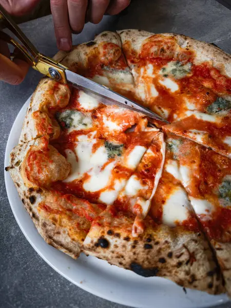 Lezzetli pizza bir tabakta makasla dilimleniyor, erimiş dört peynir ve çeşitli malzemeyi vurguluyor, eşsiz bir yemek deneyimi gösteren iştah açıcı bir görüntü yaratıyor. Boşluğu kopyala