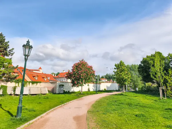Prag, Çek Cumhuriyeti - 5 Mayıs 2025 Prag, Czechia 'daki Picturesque Park, dolambaçlı bir yol, yemyeşil çimenler, çiçek açan kestane ağacı ve parçalı bulutlu gökyüzü altındaki kırmızı çatılı binalar