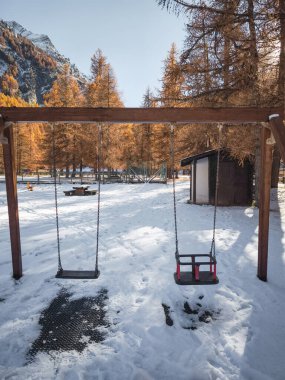Aosta, İtalya 'da sonbahar ağaçlarıyla çevrili, karlı bir oyun alanında boş salıncaklar. Arka plandaki dağlar huzurlu manzarayı güzelleştirir Frazionne Lillaz