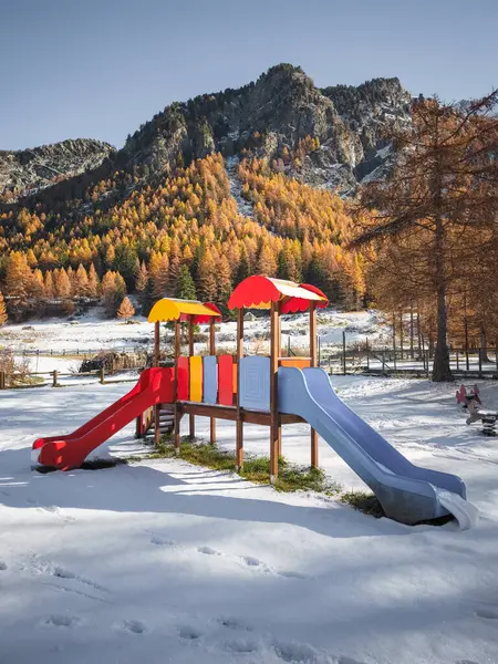 Parlak oyun alanı ekipmanları karla kaplı zemin ile tezat oluşturur, sonbahar ağaçları ve Aosta 'daki engebeli dağlara karşı kurulur, İtalya Frazionne Lillaz