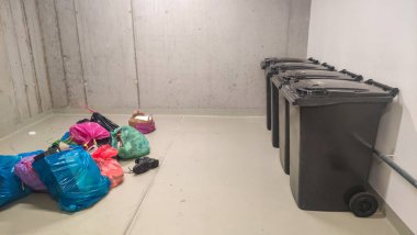 Beton bir odada, bir sıra büyük çöp tenekesinin yanında taşan çöp torbaları, uygun atık imhası ve geri dönüşüm uygulamalarının zorluklarını ve önemini vurguluyor.