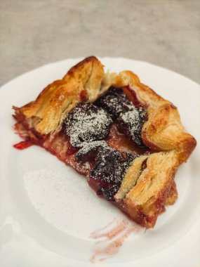 Krema şekeri serpilmiş bir dilim erik galetesinin kapağını kapatın, beyaz bir tabakta servis edilir, altın kabuğu ve sulu erik dolgusu sergilenir.