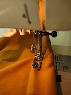 Dikiş makinesinin yakın görüntüsü iğne ve ütücü ayağın bir parça sarı kumaşı dikmesi, giysi veya tekstil oluşturma sürecini vurguluyor.