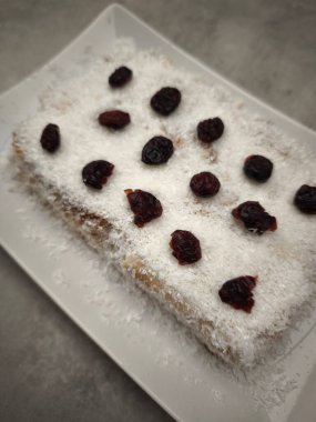 Hindistan cevizi gevreğiyle kaplanmış ve kızılcıklarla süslenmiş dikdörtgen noel pudingi aperatif olarak beyaz dikdörtgen bir tabak parti yemeği üzerinde duruyor, Yeni Yıl arifesi yemeği, Noel ya da doğum günü yemeği.