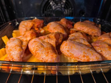 Baharatlı tavuk kanatları cam bir tabakta dilimlenmiş patates ve havuç pişiriyor, mutfağı ağız sulandırıcı aromalarla dolduruyor, ve rahatlatıcı bir yemek yaratıyor.