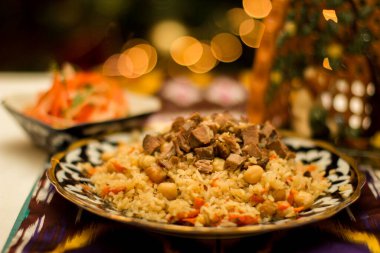 Renkli süslemelerle çevrili lezzetli et ve nohutlarla güzel bir şekilde düzenlenmiş Özbek pilav tabağı, yemek pişirme geleneği ve sıcaklığı için davetkar bir ortam yaratıyor.