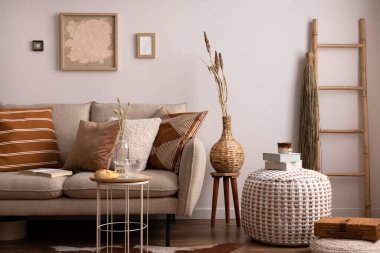 Kahverengi halı, yastık, ekose, kahve masası ve kişisel aksesuarlarla bej koltuklu oturma odasının şık kompozisyonu. Bej renkli duvar ve sahte poster. Ev dekoru. Şablon