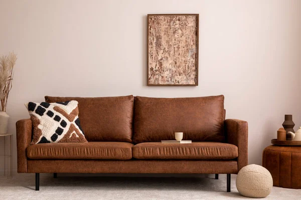 Warm and cozy interior of living room space with mock up poster fame, brown sofa, beige carpet, patterned pillow, modern pouf, vase with dried flowers, decoration and personal accessories. Cozy home decor. Template.
