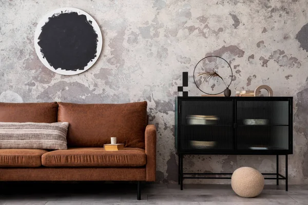 Interior design of loft industrial apartment with mock up poster frame modern brown sofa, black commode, round pillows and personal accessories. Gray concrete wall. Home decor.