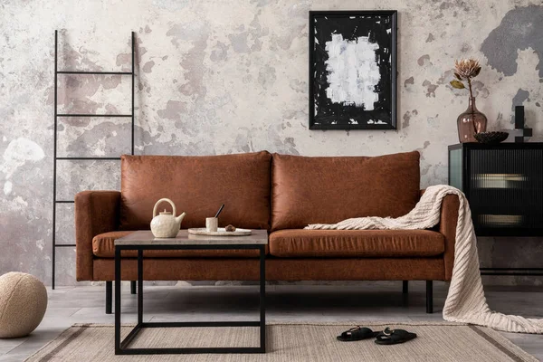 Interior design of loft industrial apartment with mock up poster frame, brown sofa, square coffee table, black commode and stylish personal accessories. Concrete gray wall. Home decor. Template.