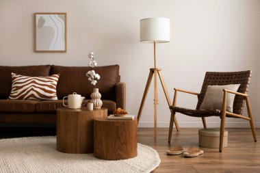 Interior design of stylish and elegant room with brown sofa, rattan armchair, wooden coffee tables, tea pot, mock up poster frame, decoration and personal accessories. Warm home decor. Template. 
