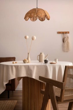 Boho and cozy interior of dining room with round wooden table, design chair, brown sofa, commode, pendant lamp, tea pot, decoration and elegant accessories. Stylish home decor. 