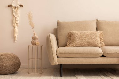 Minimalist composition of living room interior with classic beige sofa, round coffee table, vase with dried flowers, brown pouf, macrame and personal accessories. Home decor. Template.