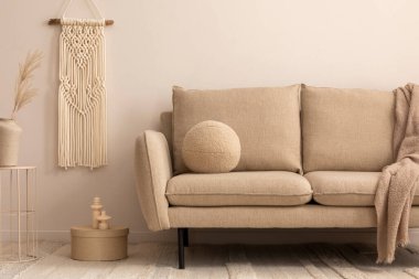Minimalist composition of living room interior with classic beige sofa, round coffee table, vase with dried flowers, brown pouf, macrame and personal accessories. Home decor. Template.