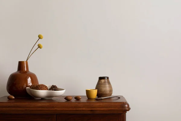 Minimalist composition of living room interior with copy space, wooden commode, stylish vase with dried flowers, bowl with coconut , nuts, beige wall and personal accessories. Home decor. Template.