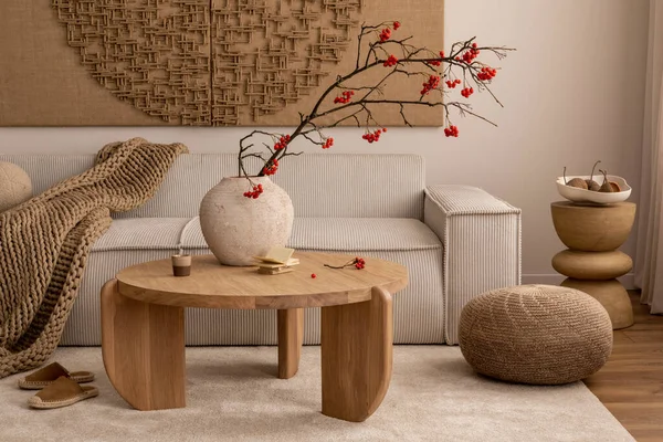 Creative composition of living room interior with wall art paint, modern beige sofa, round wooden coffee table, vase with rowan, brown pouf, slippers and personal accessories. Home decor. Template.