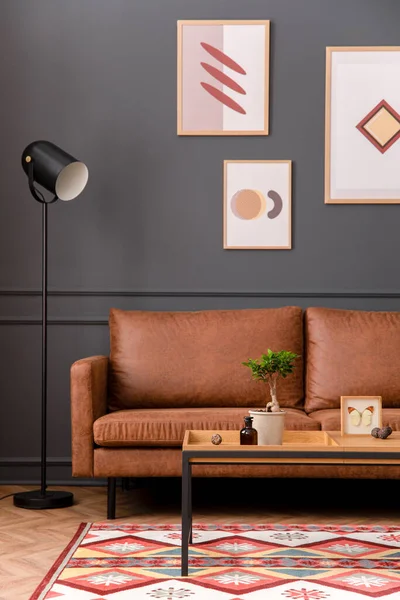 Aesthetic interior of living room with mock up poster frame, brown sofa, wooden coffee table, black lamp, patterned rug, gray wall, plants in flowerpot and personal accessories. Home decor. Template