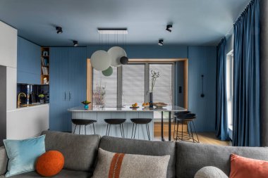 Interior design of kitchen interior with marble kitchen island, black chokers, blue kitchen furnitures, modern lamp, window, gray sofa, colorful pillows and personal accessories. Home decor. Template.
