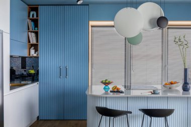 Creative composition of kitchen interior with marble kitchen island, blue kitchen furnitures, black chokers, modern lamp, bowl with fruits, big window and personal accessories. Home decor. Template. 