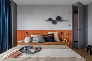 Interior design of elegant bedroom with big orange bed, beige and grey bedclothes, blue curtain, modern lamp, night stand, vase with dried flowers and personal accessories. Home decor. Template.