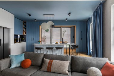 Interior design of kitchen interior with marble kitchen island, black chokers, blue kitchen furnitures, modern lamp, window, gray sofa, colorful pillows and personal accessories. Home decor. Template.