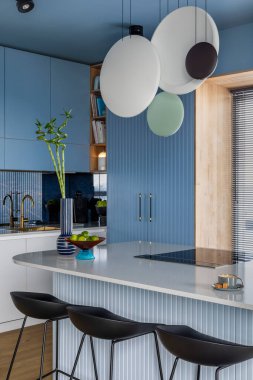 Interior design of kitchen interior with marble kitchen island, blue wall, black chokers, bowl with fruits, big window, cup, wooden floor, lamp on wall and personal accessories. Home decor. Template.