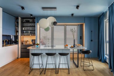 Interior design of kitchen space with marble island, black chockers, modern lamp, wooden wall, blue kitchen furnitures, vase with dried flowers and personal accessories. Home decor. Template. 