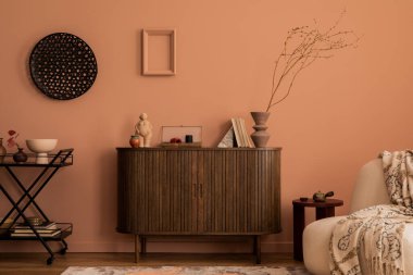 Warm and cozy living room interior with wooden commode, stylish armchair, patterned plaid, vase with dried flowers, sculpture, ornament on the wall and personal accessories. Home decor. Template. 