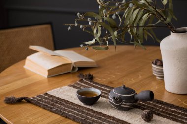 Creative composition of living room interior with wooden round table, book, beige vase with leaves, tea in cup, pitcher, chair, patterned table pad and personal accessories. Home decor. Template.