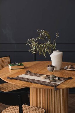 Interior design of aesthetic and elegant room with round wooden table, vase with dried flowers, book, bowl with nuts, rattan chairs, dark wall, incense and personal accessories. Home decor. Template.